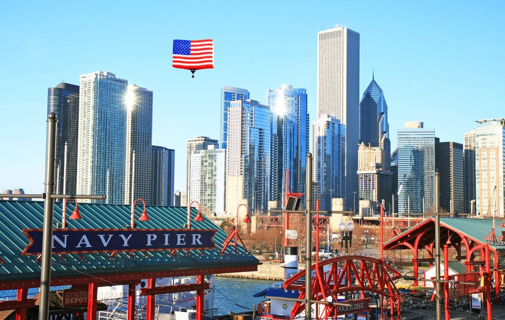 Chicago skyline with modern skyscrapers, the Navy Pier sign in the foreground, and a large American flag suspended beneath a helicopter in the blue sky.
