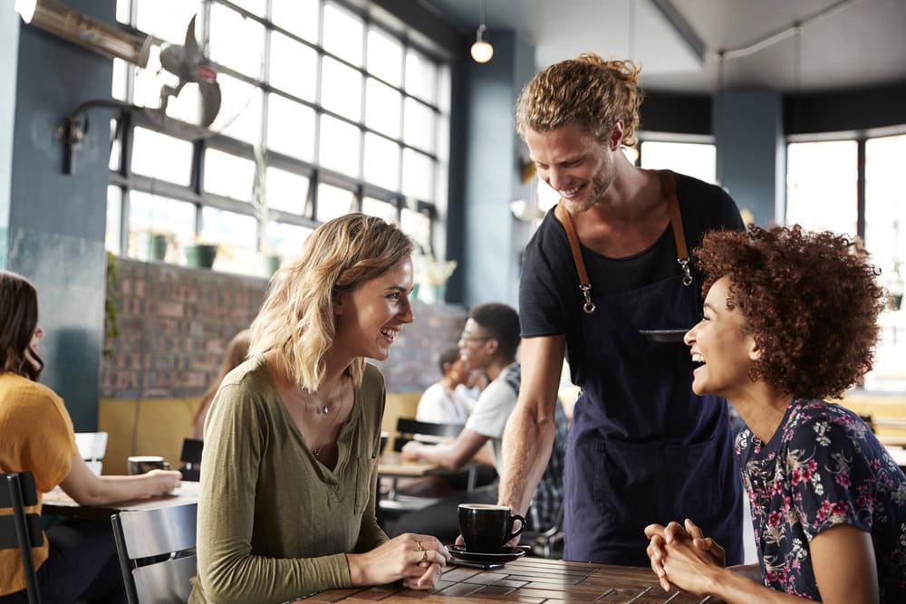 A server hands a cup of coffee to two women sitting at a table, smiling and talking in a bright, modern cafe.