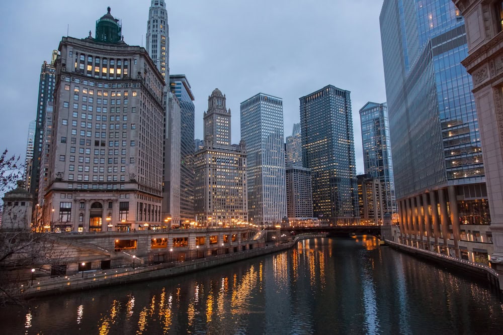 City skyline with tall buildings reflecting in a river at dusk, with street lights illuminating the area and a cloudy sky overhead.