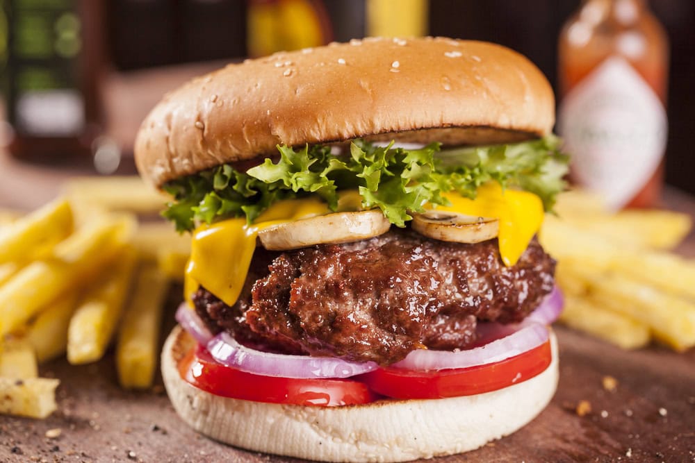 A cheeseburger with lettuce, tomato, onions, pickles, mushrooms, and cheddar cheese, served with a side of French fries.