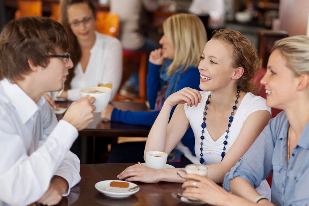 Three people sitting at a café table, drinking coffee and talking, with others seated in the background.