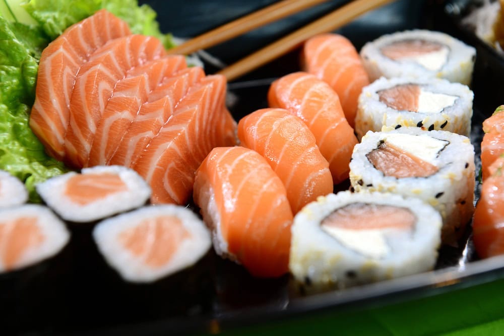 A close-up of assorted sushi including salmon sashimi, salmon nigiri, and various salmon rolls, served on a black tray with lettuce garnish and chopsticks in the background.