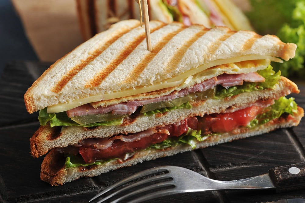A toasted club sandwich with layers of ham, cheese, lettuce, tomato, and bacon, secured with a toothpick, on a dark wooden board with a fork nearby.