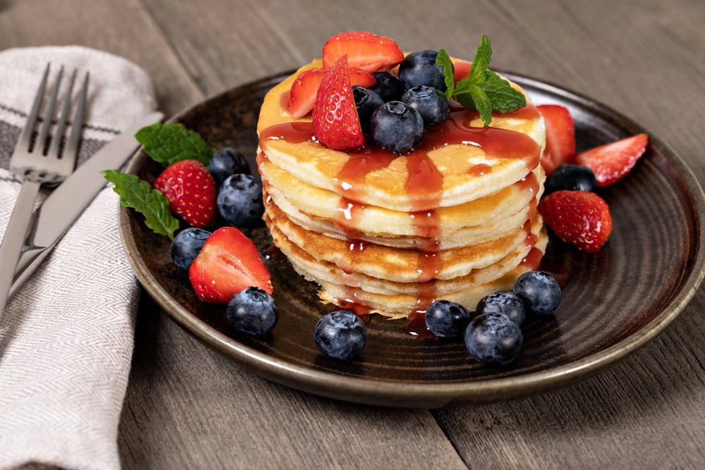 A stack of pancakes topped with sliced strawberries, blueberries, mint leaves, and syrup on a brown plate, with more berries scattered around. A fork and knife rest on a napkin beside the plate.