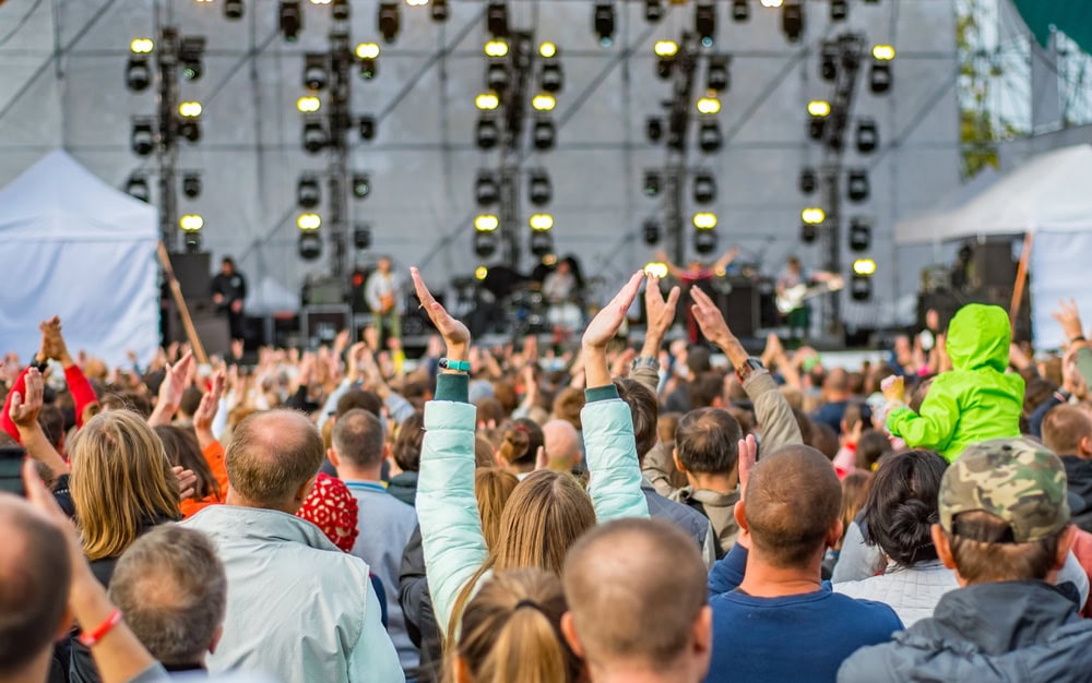 A large crowd facing a stage with bright lights, watching a live outdoor music performance during the day.