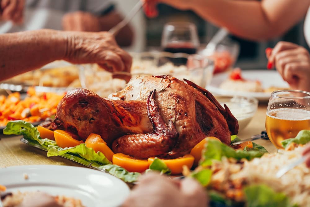 A roasted turkey garnished with vegetables is served on a table, surrounded by people reaching for food and drinks.