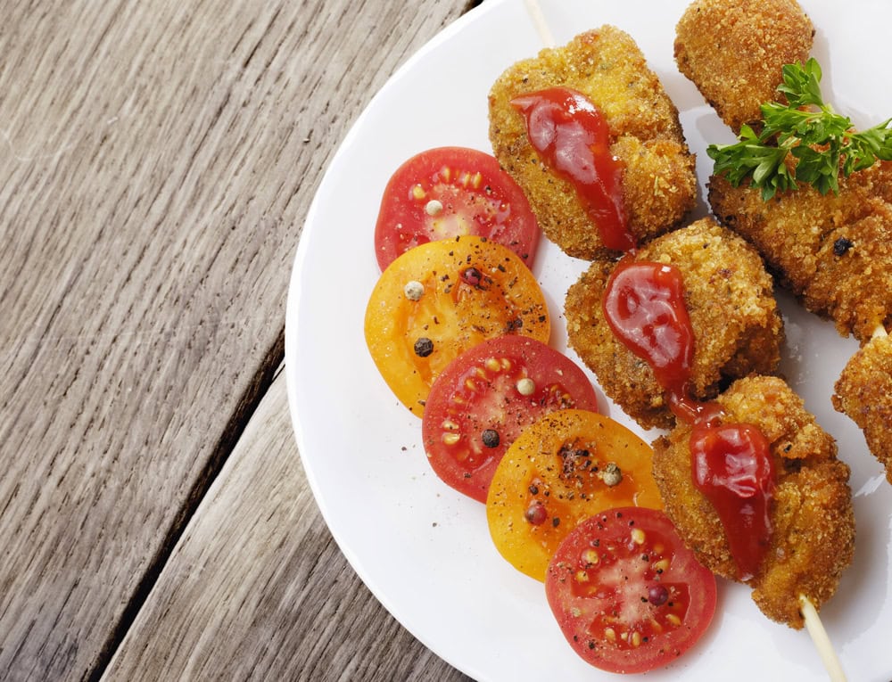 A plate with breaded skewered meat topped with ketchup, garnished with parsley, and sliced red and yellow tomatoes seasoned with pepper.