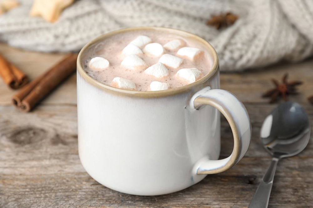 A white mug filled with hot chocolate topped with mini marshmallows sits on a wooden surface beside a spoon, cinnamon sticks, and a knitted fabric.