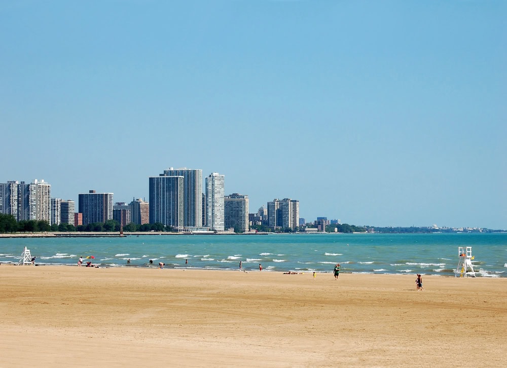 Sandy beach with a few people and lifeguard chairs, calm water, and a city skyline with high-rise buildings under a clear blue sky.