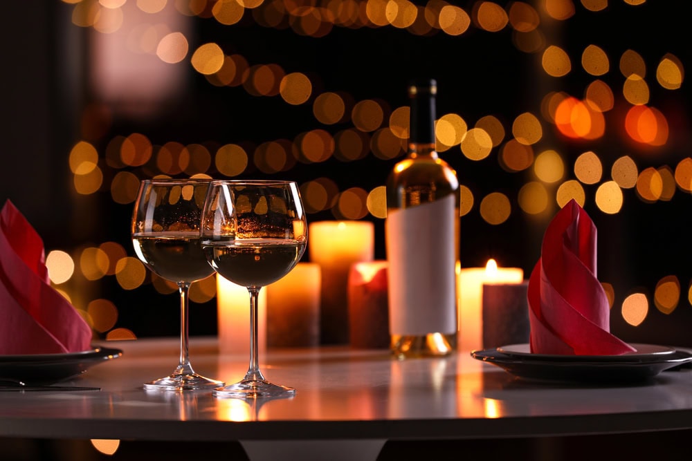 Two glasses of wine, a wine bottle, candles, and folded red napkins on a table with blurred warm lights in the background.