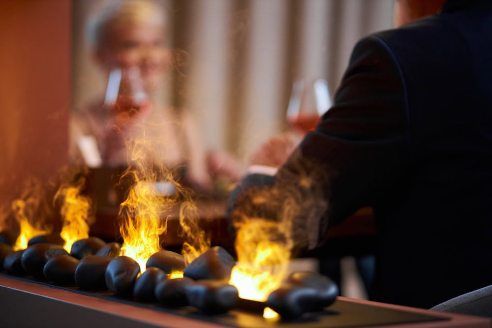 Two people sit at a table with glasses of wine, separated by a decorative row of black stones with visible flames.
