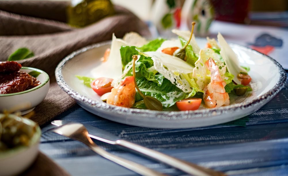 A plate of fresh salad with shrimp, cherry tomatoes, lettuce, cheese shavings, and capers, served on a white dish beside a fork and knife.