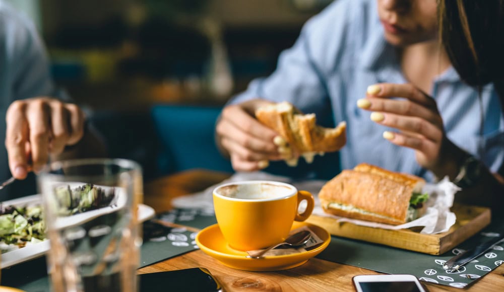 Two people are seated at a table eating sandwiches; one holds a sandwich, while a yellow coffee cup and a plate with salad are visible on the table.