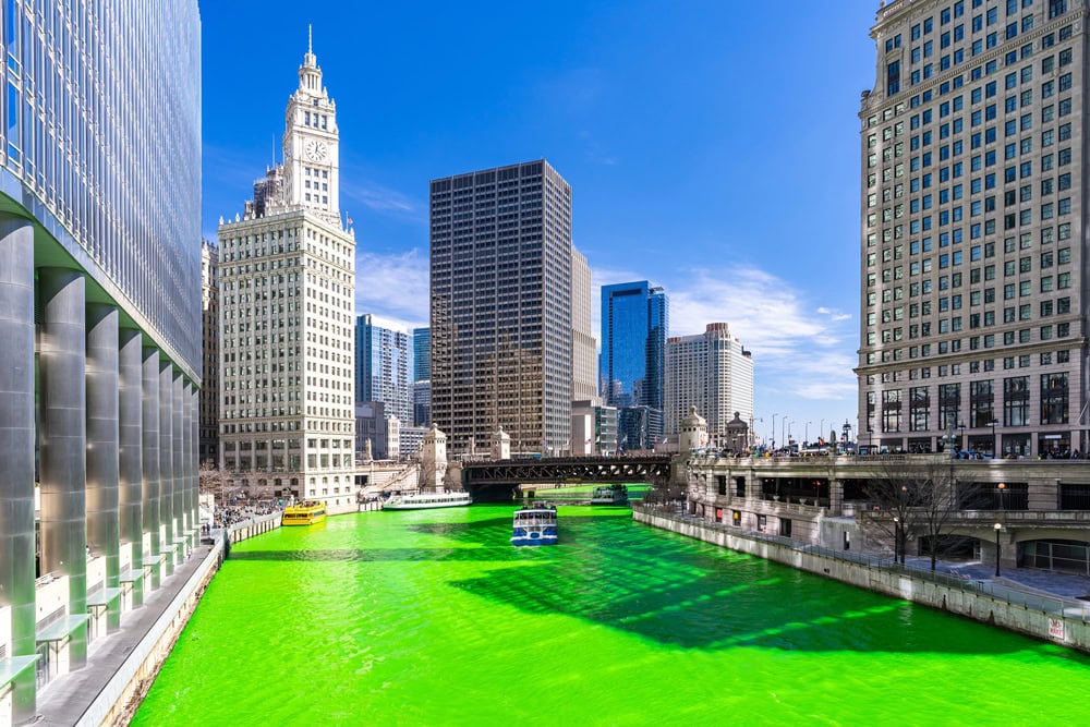 A city river dyed bright green flows between tall buildings under a clear blue sky, with several boats and a bridge visible.