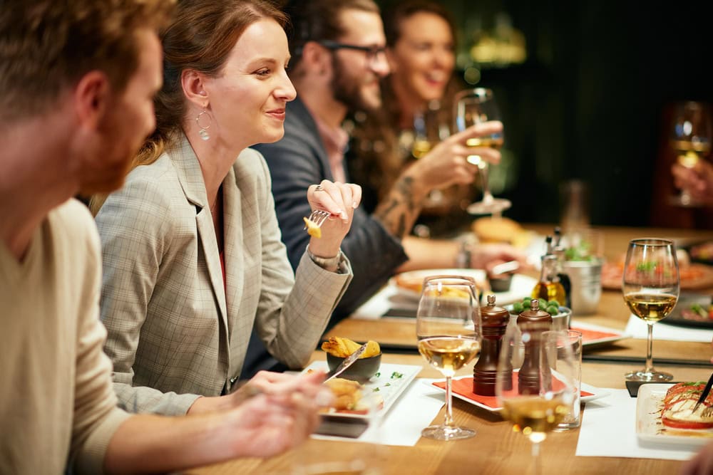 A group of people sit at a restaurant table, eating food and drinking wine, with plates, glasses, and condiments visible on the table.