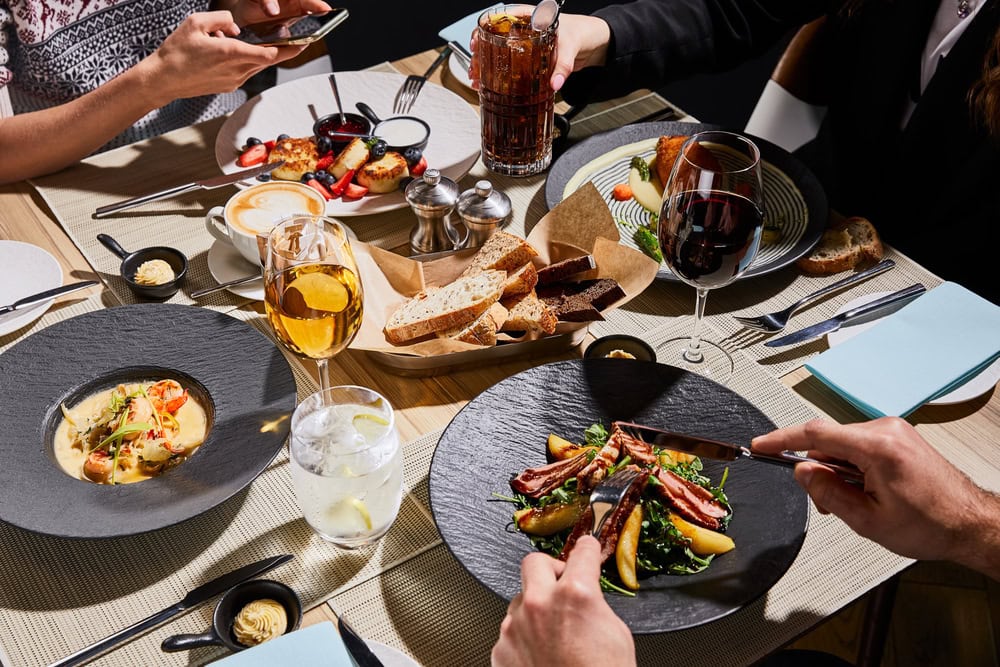 Three people dining at a restaurant table with plated gourmet food, drinks including wine and soda, bread basket, and desserts; one person is using a smartphone.