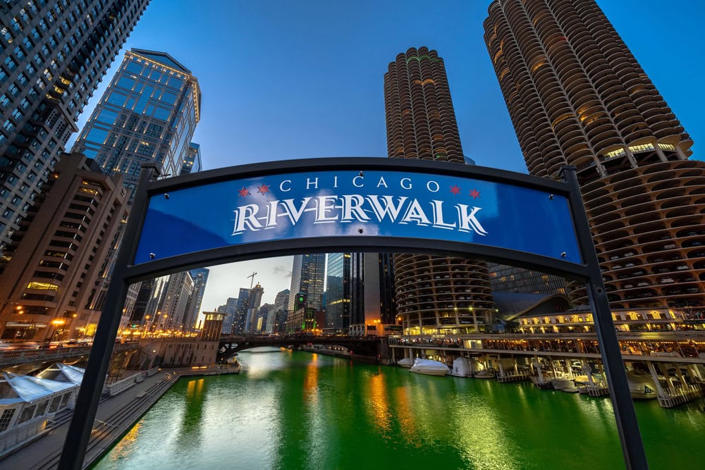 Chicago Riverwalk sign with tall buildings and the green Chicago River in the background at dusk.