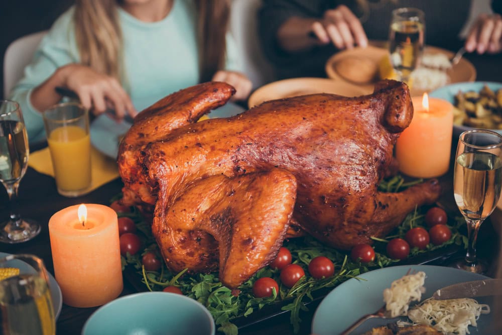 A roasted turkey is served on a platter with cherry tomatoes and greens, surrounded by candles, drinks, and people at a dining table.