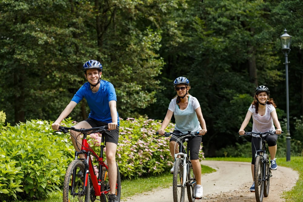 Three people wearing helmets ride bicycles on a dirt path through a green, wooded park, with trees and bushes lining the trail.