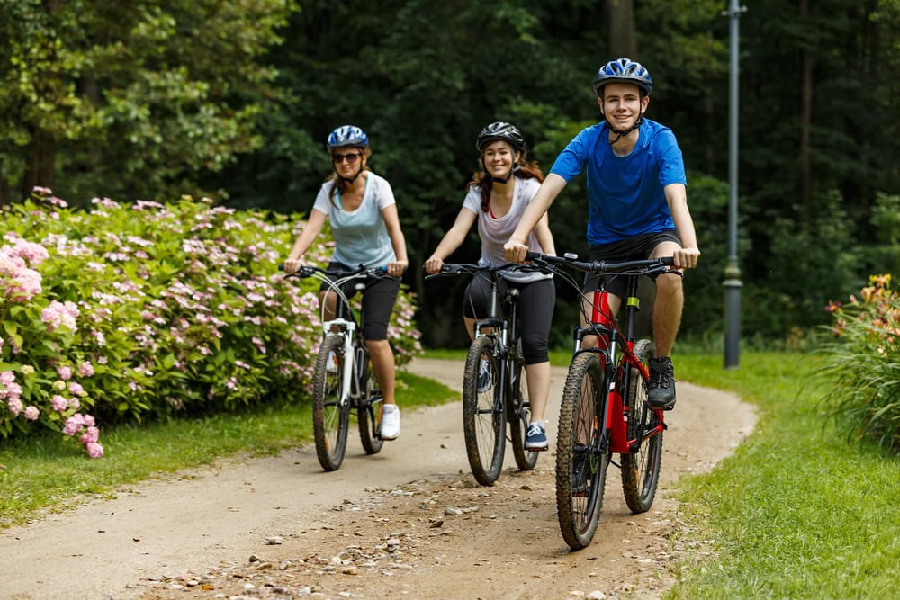 Three people wearing helmets ride bicycles along a dirt path in a park with green trees and blooming flowers.