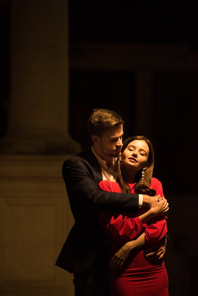 A man in a suit hugs a woman in a red dress from behind as they stand together in dim lighting.