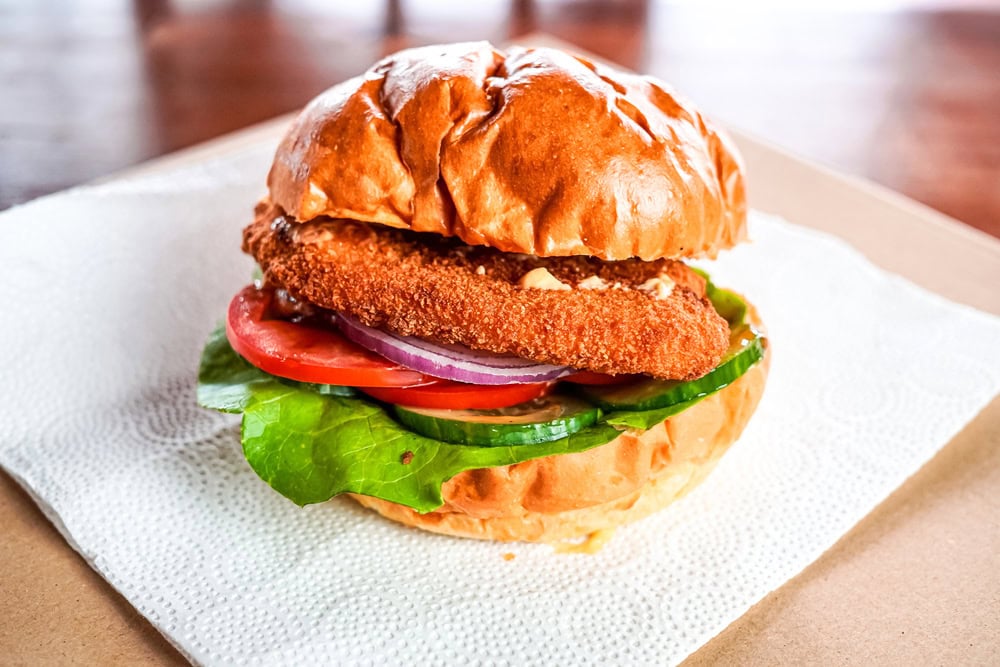 A breaded fried fish sandwich with lettuce, tomato, cucumber, and red onion on a bun, placed on a white paper napkin.