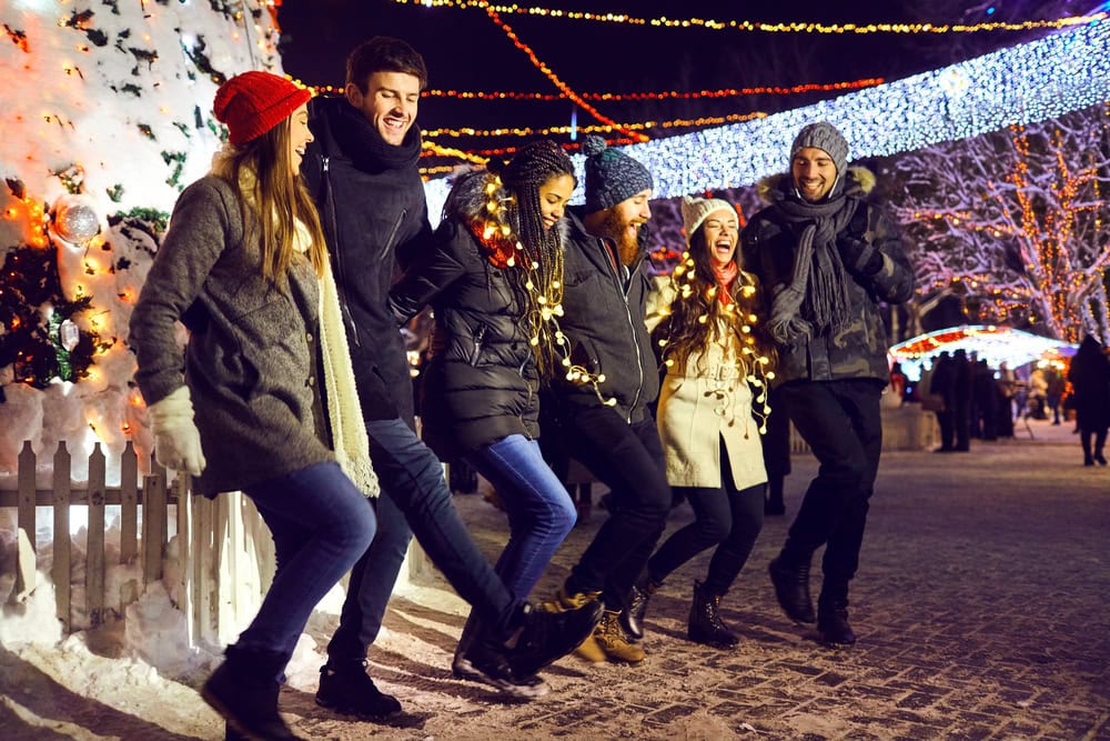 Six people in winter clothing stand in a row, linking arms and kicking their legs, celebrating at an outdoor holiday event with festive lights and a decorated tree.