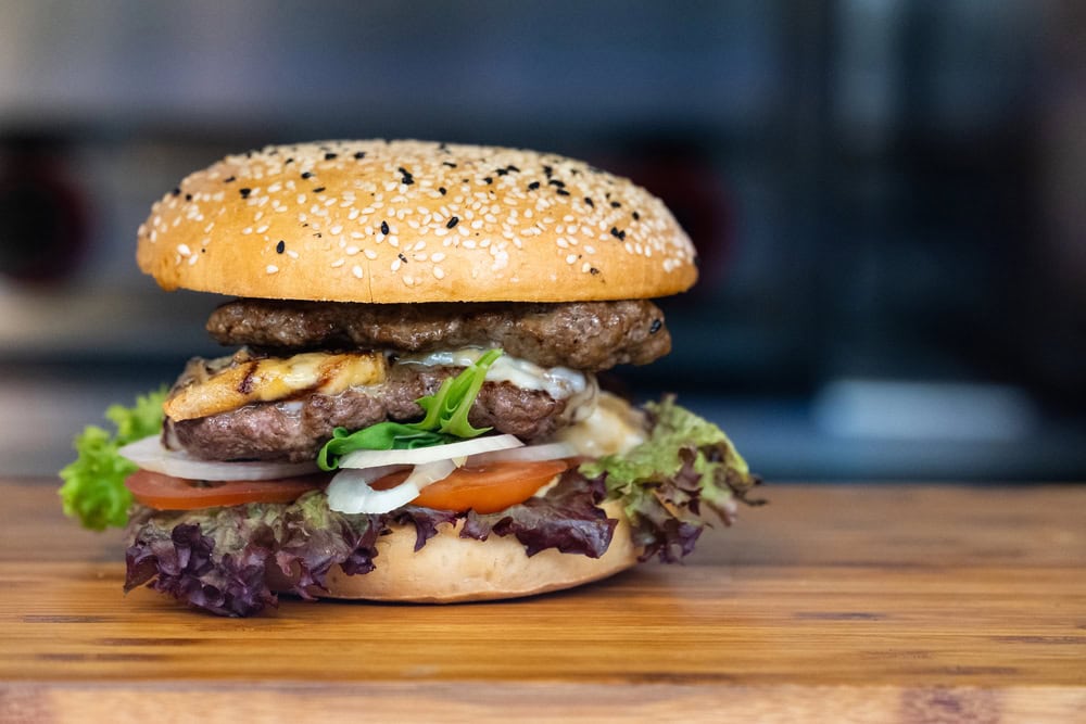 A sesame seed hamburger bun with two beef patties, cheese, lettuce, tomato, and onion sits on a wooden surface.