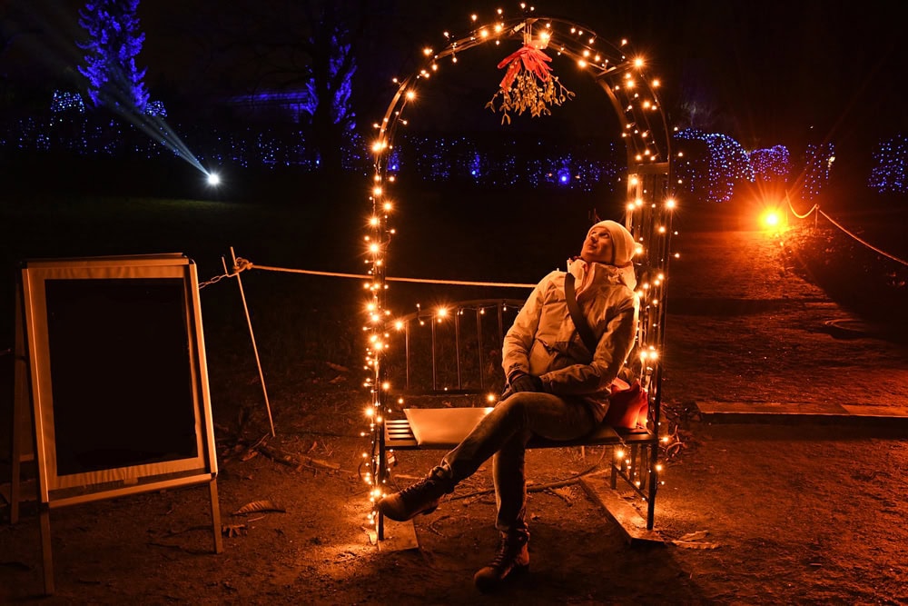 A person sits on a bench decorated with string lights at night, looking up. The area around is dark with blue and orange lights in the background.