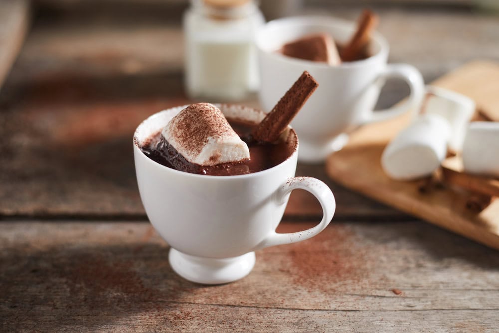 A white cup of hot chocolate topped with marshmallows and a cinnamon stick sits on a wooden table, with another cup, marshmallows, and a jar in the background.