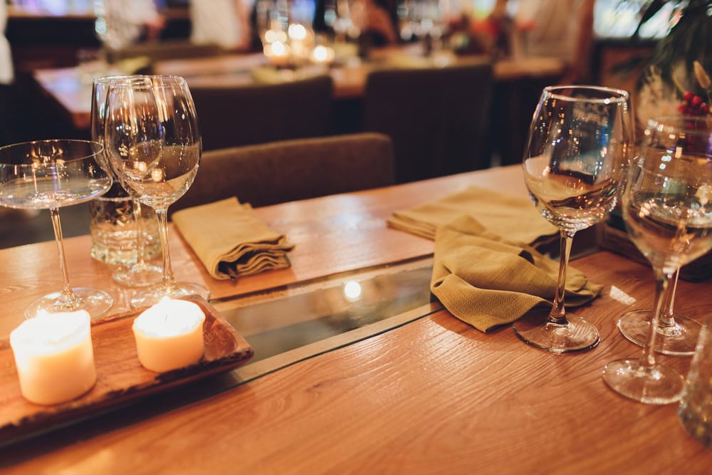 A wooden restaurant table set with wine glasses, folded napkins, and lit candles, with blurred diners and lights in the background.