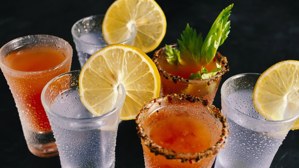Six drinks, three clear and three red, are garnished with lemon slices; the red drinks have spiced rims and celery, while the clear drinks are served in frosted glasses.
