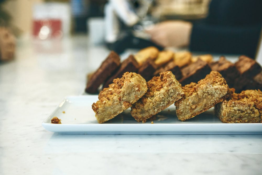 A close-up of a white rectangular plate holding crumbly dessert bars, displayed on a marble counter with blurred baked goods in the background.