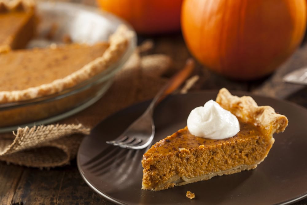 A slice of pumpkin pie with a dollop of whipped cream on a brown plate, next to a fork, with a whole pie and pumpkins in the background.