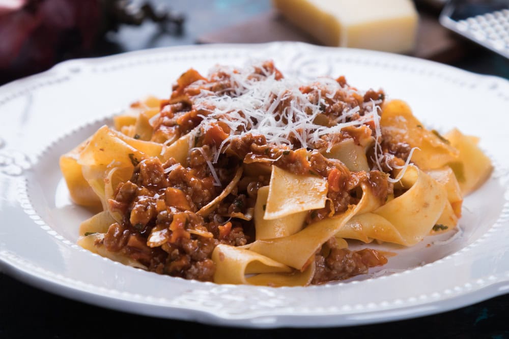 A plate of pappardelle pasta topped with meat sauce and grated cheese, served on a white decorative plate.