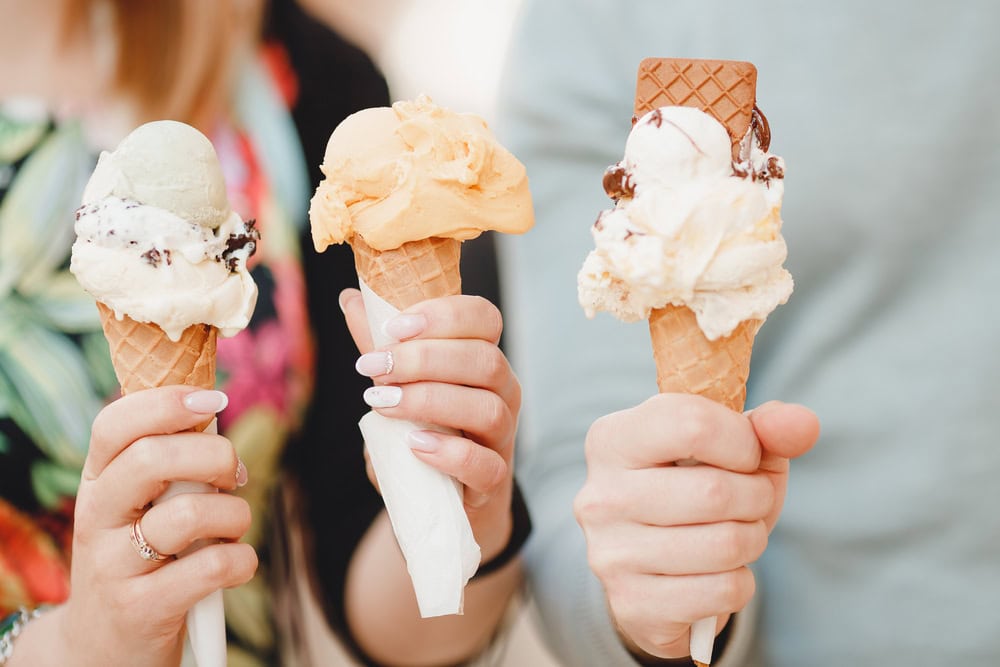 Three people hold ice cream cones with various flavors and toppings, including chocolate pieces and wafer, against a blurred background.