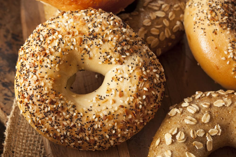 Close-up of an everything bagel with visible sesame, poppy seeds, and seasoning, next to other bagels with different toppings.
