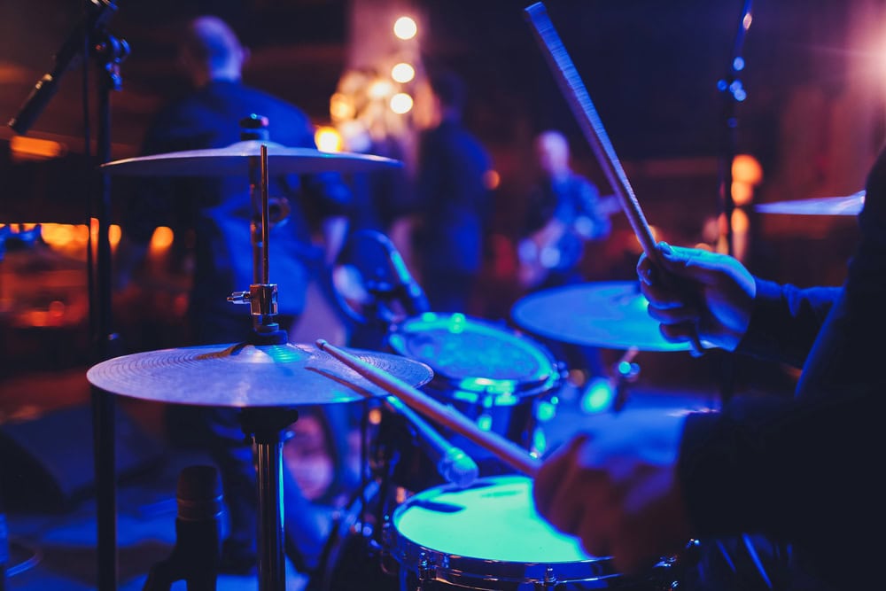 Close-up of a drummer playing a drum set on stage with blurred band members and colorful lights in the background.
