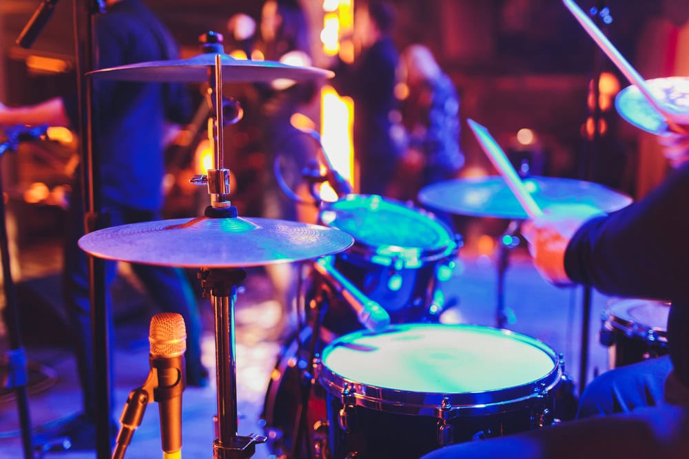 A close-up view of a drum set with a microphone in front, while musicians perform on a dimly lit stage in the background.