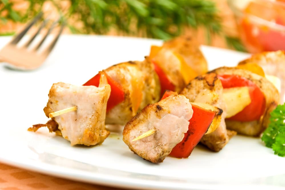 Close-up of grilled chicken and vegetable skewers on a white plate, with red peppers and herbs visible in the background.