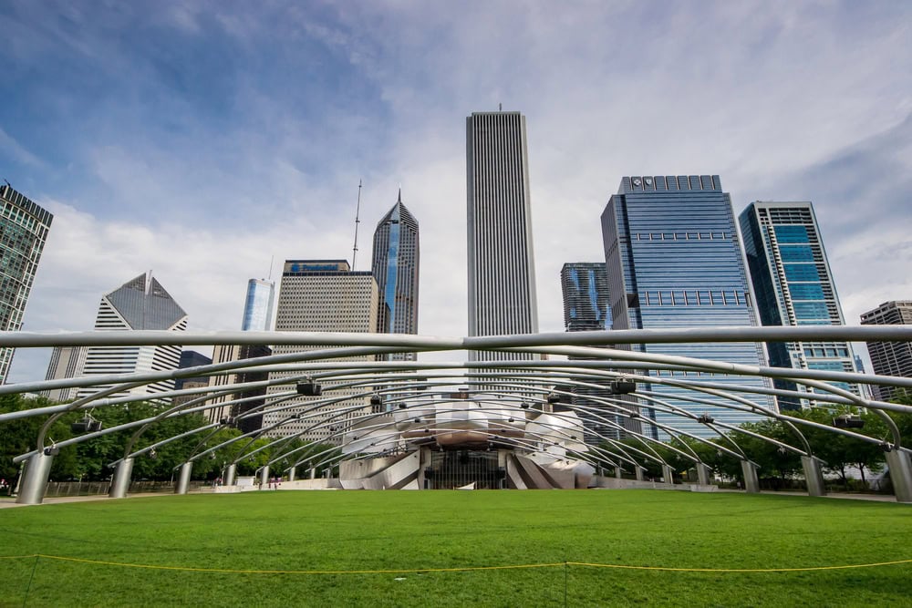 A view of Chicago’s city skyline behind the Jay Pritzker Pavilion and its open grass area in Millennium Park, under a partly cloudy sky.
