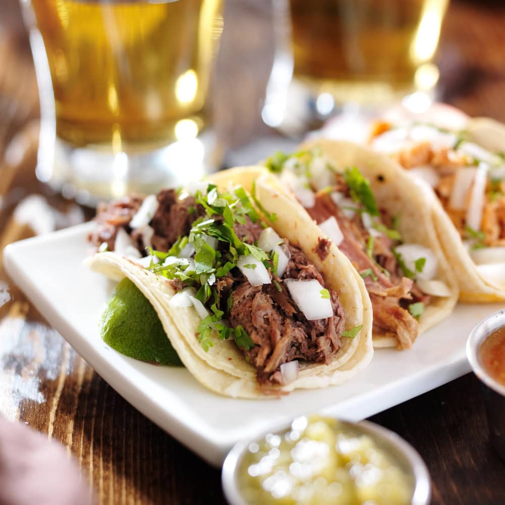 Three soft tacos filled with shredded meat, diced onions, and chopped cilantro are served on a white plate with a lime wedge, with drinks and salsa in the background.