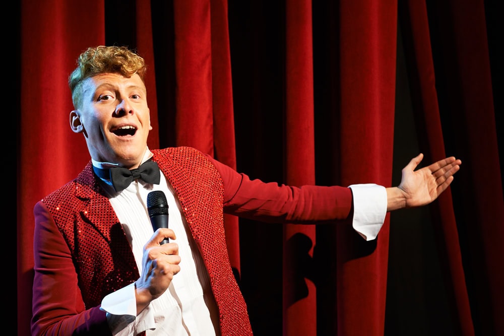 A person in a red tuxedo jacket and bow tie holds a microphone and gestures toward red stage curtains.