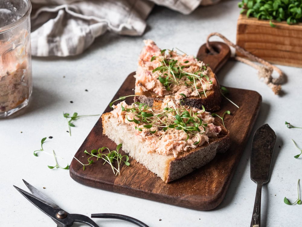 Two slices of rustic bread topped with a pink spread and fresh microgreens sit on a wooden board, with kitchen shears and a glass jar nearby.