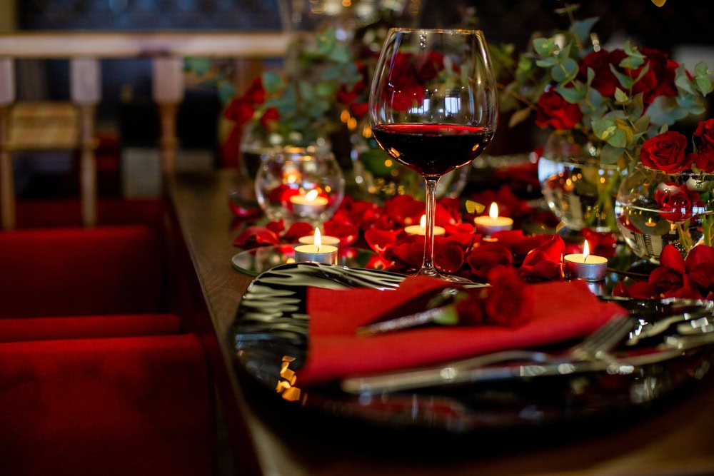 A table set with a glass of red wine, red napkins, silverware, rose petals, candles, and floral arrangements in a dimly lit, elegant setting.