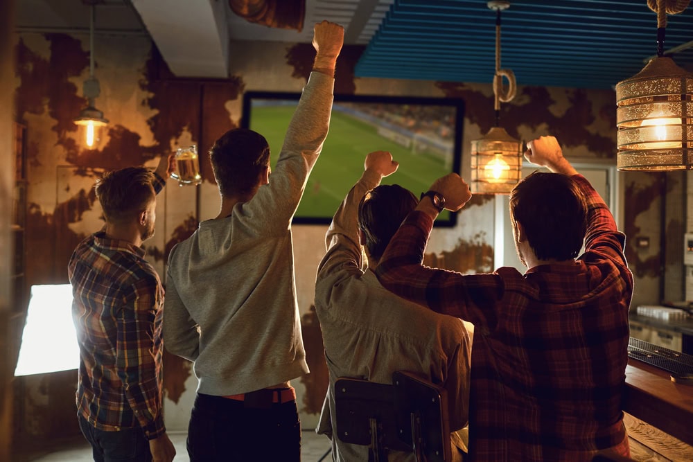 Four people stand and raise their arms while watching a soccer match on a TV in a dimly lit bar with rustic decor.