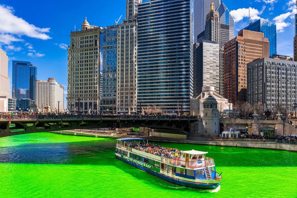 A tour boat travels on the Chicago River, which is dyed bright green for St. Patrick’s Day, with tall city buildings in the background.