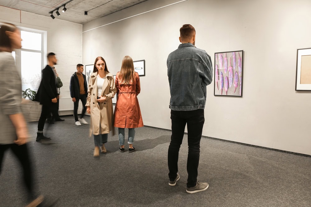 People viewing artwork in a modern gallery; some are closely observing the pieces, while others walk or stand in groups in a minimalist, well-lit space.