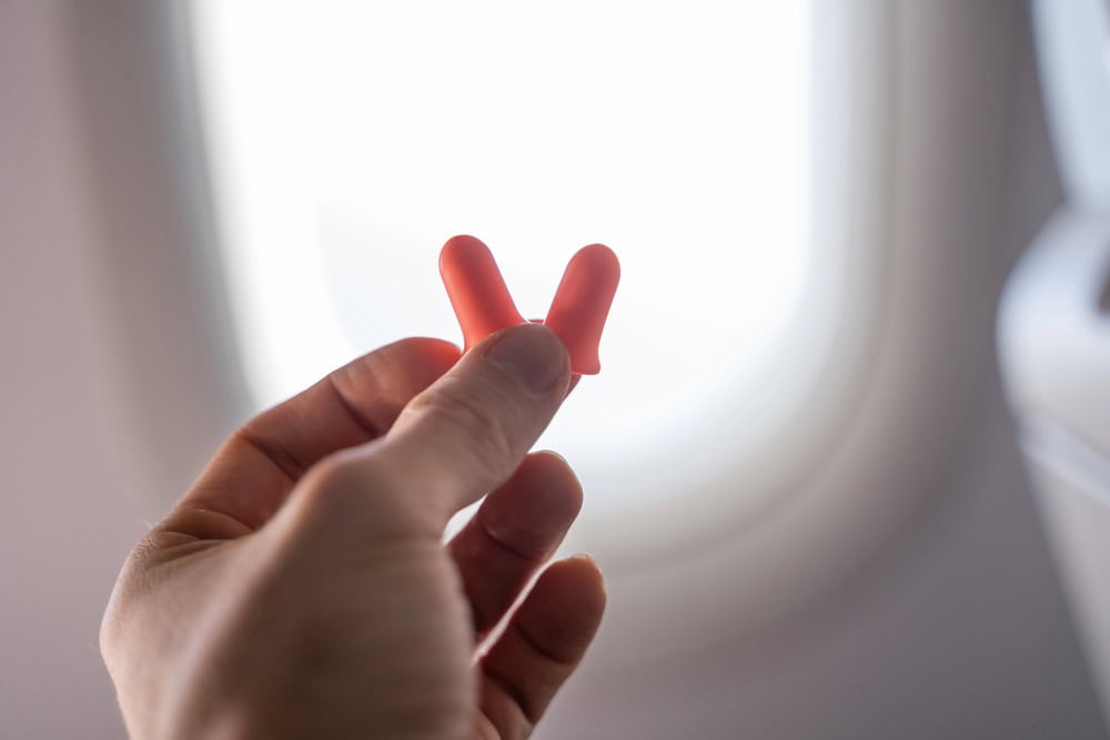 A hand holding two orange earplugs in front of an airplane window.
