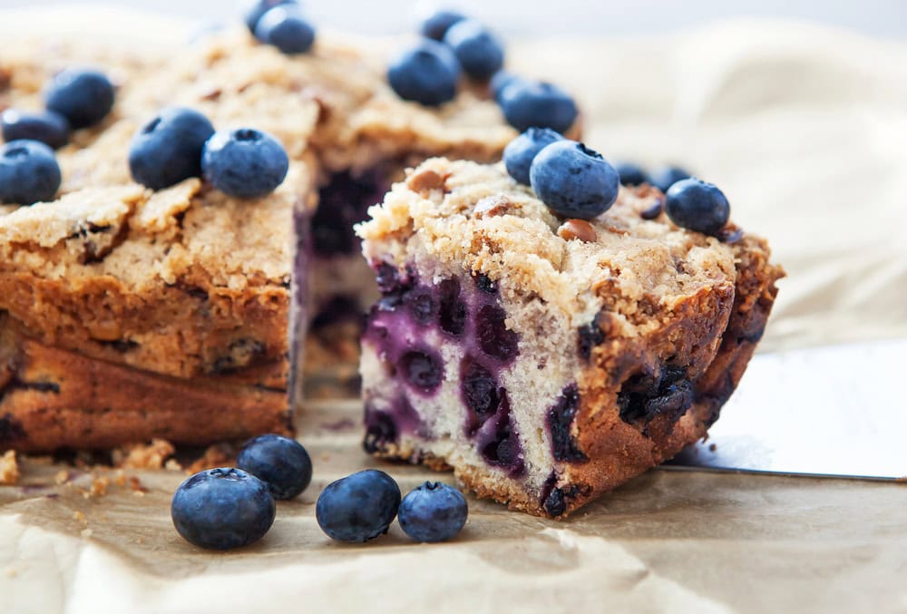 A slice of blueberry cake with a crumb topping is being served, showing blueberries inside and on top of the cake.
